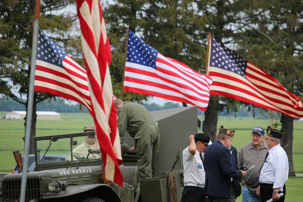 pexels-photo-8253834-8253834 Group of veterans and soldiers by military vehicle with American flags in the background, symbolizing patriotism.